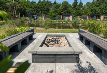 Udendørs ildsted med moderne bænke ved en dam, omgivet af grønne planter og feriehytter i Holland.