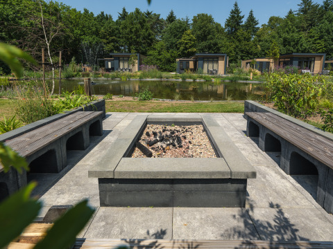 Moderne Feuerstelle mit Sitzbänken am Teich, umgeben von Pflanzen und Lodges im Veluwepark de Bosgraaf.