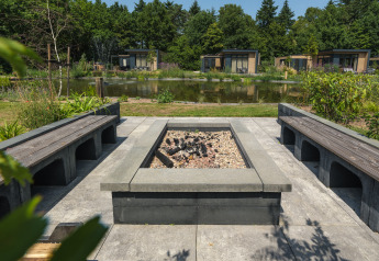 Udendørs ildsted med moderne bænke ved en dam, omgivet af grønne planter og feriehytter i Holland.