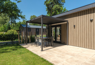 Udendørs terrasse ved en moderne lodge med have, bord og stole under en pergola på en solrig dag.