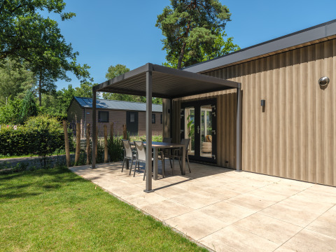 Patio exterior en una moderna casa de campo con jardín, mesa y sillas bajo una pérgola en un día soleado.
