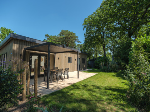 Udendørsbillede af en moderne lodge med overdækket terrasse, havemøbler og grøn have under blå himmel.