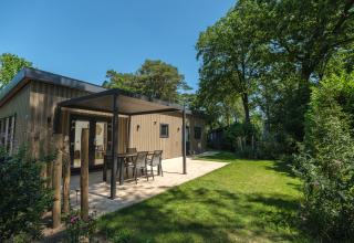 Udendørsbillede af en moderne lodge med overdækket terrasse, havemøbler og grøn have under blå himmel.