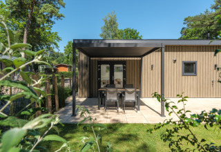 Cabaña moderna de madera con terraza cubierta, comedor exterior y jardín rodeado de árboles verdes.
