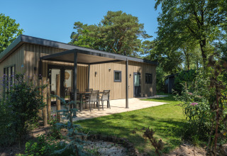 Moderne lodge med træfacade, overdækket terrasse og havemøbler, omgivet af grøn natur og træer.