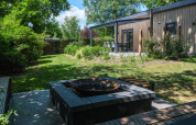 Vue du jardin ensoleillé sur la lodge Loevestein Deluxe, terrasse, façade bois et coin brasero moderne.