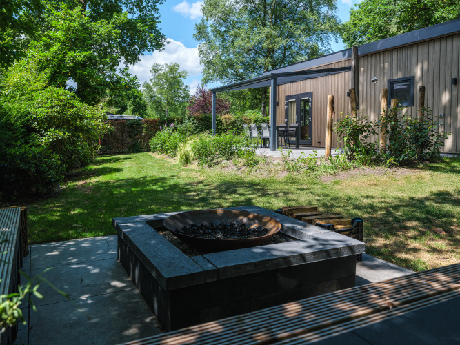 Blick auf die Loevestein Deluxe Lodge mit moderner Holzfassade, Garten und Feuerstelle an einem sonnigen Tag.