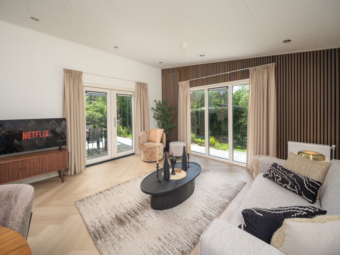 Modernes Lodge-Wohnzimmer mit großen Fenstern, Holzvertäfelung, beigem Sofa und Netflix auf dem Fernseher.