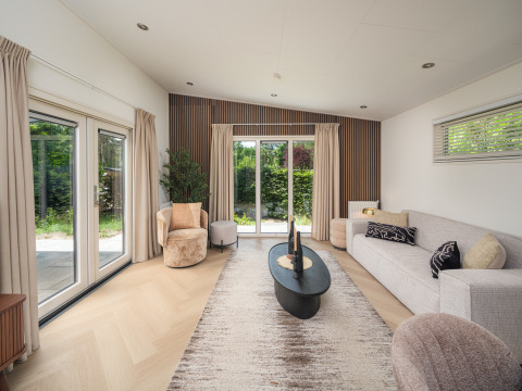 Moderne opholdsstue i en lodge med store vinduer, neutral indretning, beige gardiner og grøn udsigt.