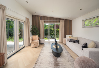 Moderne opholdsstue i en lodge med store vinduer, neutral indretning, beige gardiner og grøn udsigt.