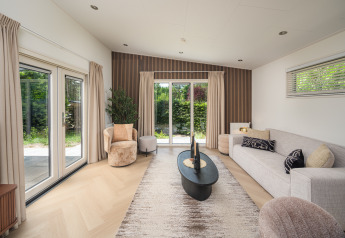 Modern lodge living room with large windows, neutral decor, beige curtains, and a view of green nature.