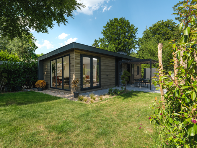 Lodge moderno Zypendaal con ventanas grandes, jardín cuidado y terraza, rodeado de árboles y arbustos verdes.