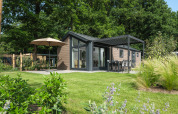Moderne Lodge mit großer Terrasse, Gartenmöbeln und Sonnenschirm in einer grünen, waldreichen Umgebung.