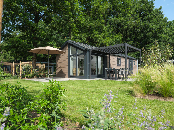 Moderne lodge omgivet af grøn have med terrasse, havemøbler og parasol midt i naturskønne omgivelser.