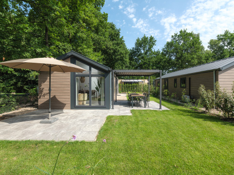 Lodge moderne avec grande terrasse, parasol et jardin, entouré d’arbres verts sous un ciel bleu.