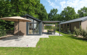 Modern lodge with a large patio, sun umbrella, and garden, surrounded by lush trees under a blue sky.
