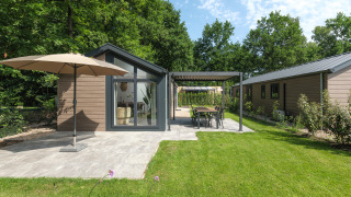 Moderne Lodge mit Terrasse, Sonnenschirm und Garten, umgeben von grünen Bäumen und blauen Himmel.