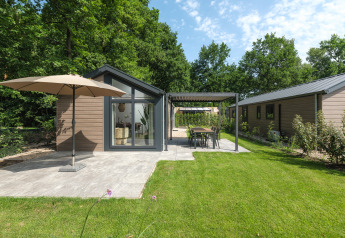 Lodge moderne avec grande terrasse, parasol et jardin, entouré d’arbres verts sous un ciel bleu.