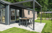 Modern lodge with outdoor dining area under a pergola on a patio, surrounded by garden and trees.