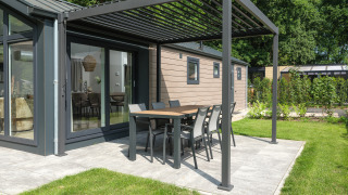 Moderne lodge met buiteneethoek onder een pergola op het terras, omgeven door tuin en groene bomen.