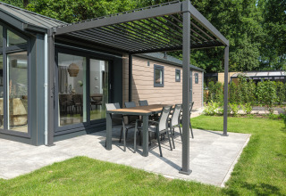 Lodge moderne avec espace repas extérieur sous pergola sur terrasse, entouré de jardin et d’arbres verts.