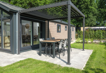 Modern lodge with outdoor dining area under a pergola on a patio, surrounded by garden and trees.