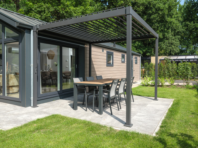 Lodge moderno con zona pranzo esterna sotto una pergola su patio, circondato da giardino e alberi verdi.