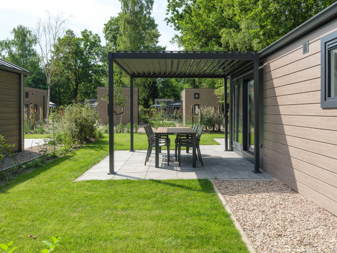 Udendørs terrasse ved en lodge med moderne overdækning, bord og stole omgivet af natur og grønne planter.