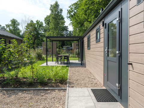 Buitenruimte bij een lodge met tuin, grindpaden en een overdekt terras met tuinstoelen onder bomen.