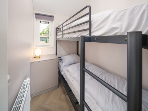 Smal lodgekamer met een metalen stapelbed, raam, radiator en een kastje met een lamp erop.