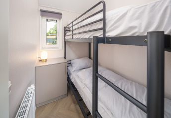 Chambre étroite de lodge avec lits superposés en métal, fenêtre, radiateur et petit meuble avec lampe.