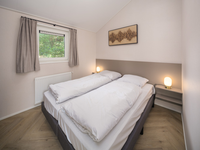 Lyst og moderne soveværelse på et lodge med dobbeltseng, store dyner, lamper og vindue med gardiner.