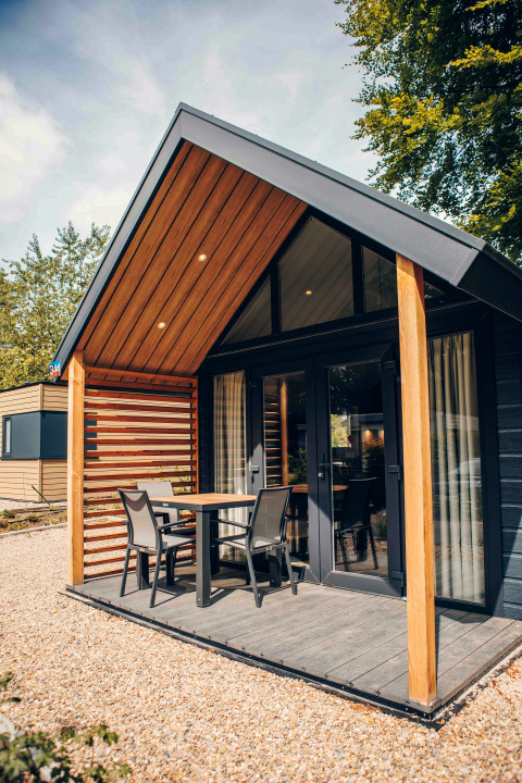 Modern tiny house with wood accents, patio set on porch, large windows, gravel yard and leafy surroundings.