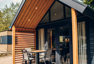 Modern tiny house with wood accents, patio set on porch, large windows, gravel yard and leafy surroundings.