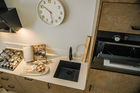 Compact tiny house kitchen with gas stove, black sink, open cookbook, and a wall clock above the counter.