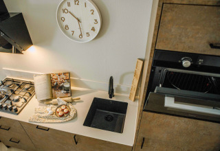 Cuisine compacte de tiny house avec évier noir, four, livre de recettes ouvert et pendule murale moderne.