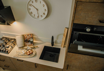 Compact tiny house kitchen with gas stove, black sink, open cookbook, and a wall clock above the counter.