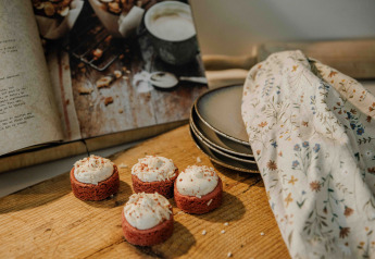 Four mini frosted cakes on a wooden board beside an open cookbook, stacked plates, and a tea towel.
