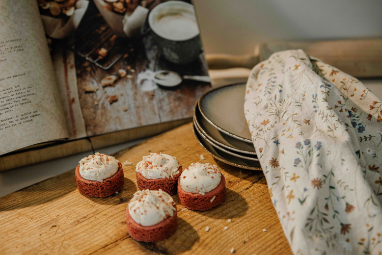 Vier kleine Kuchen mit Zuckerguss auf einem Holzbrett neben einem aufgeschlagenen Kochbuch und Tellern.