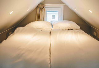 Cozy sleeping loft in a tiny house with a double bed, soft lighting, and a small window at the end.