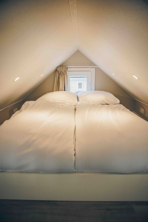 Acogedor altillo con cama doble en una tiny house, techo bajo, luz tenue y pequeña ventana al fondo.