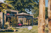 Außenbereich einer Lodge mit modernen Terrassen, Sonnenschirmen und viel Grün unter großen Bäumen.