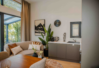 Moderne lodge-stue med sofa, planter, lille køkken, kaffemaskine, kunst og store vinduer til naturen.