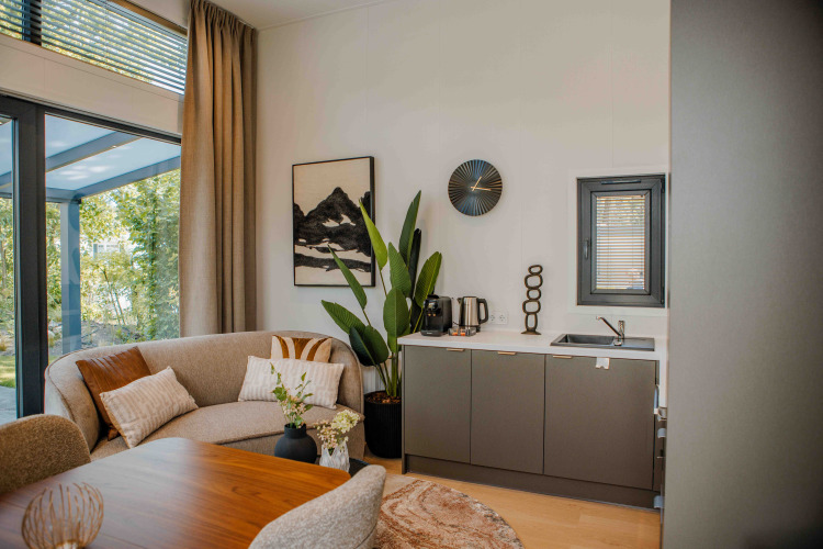 Moderne lodge met comfortabele bank, kitchenette, planten en grote ramen met uitzicht op de natuur.