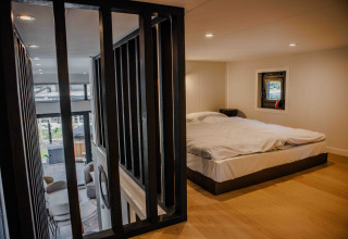 Lodge-slaapkamer met modern interieur, tweepersoonsbed, houten vloer en zicht op de woonkamer beneden.