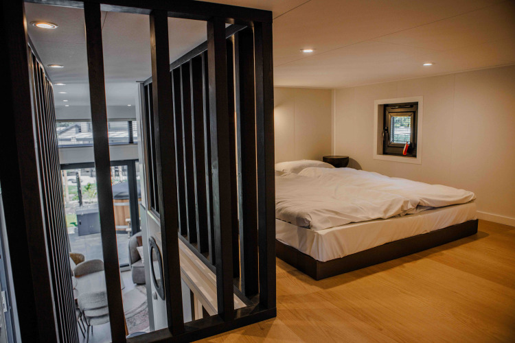 Modern lodge bedroom with a double bed, wood flooring, and a view of the living area through black slats.