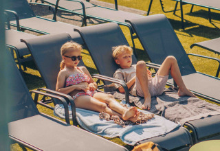 Twee kinderen ontspannen op ligstoelen met handdoeken op een zonnige dag bij een glampingaccommodatie.