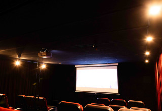 Sala de cine oscura con butacas rojas vacías, pantalla iluminada y luces suaves en un glamping.