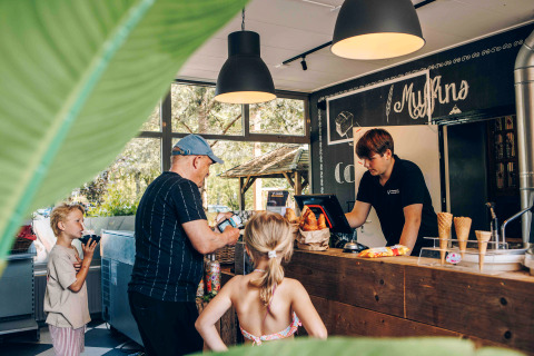 Family orders ice cream and pastries at a cozy glamping café counter, surrounded by leafy greenery outside.