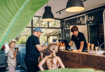 Familia compra helados y bollería en una acogedora cafetería de glamping con vegetación visible afuera.